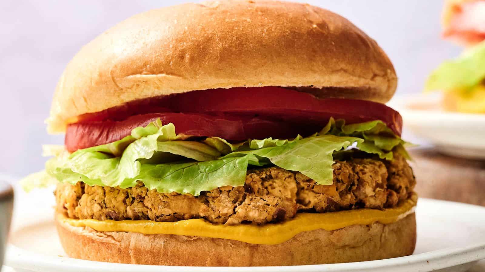 A veggie burger topped with lettuce, tomato slices, and mustard in a sandwich bun, served on a white plate.