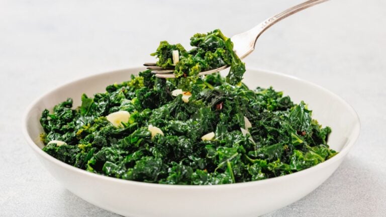A white bowl filled with sautéed kale and sliced garlic, with a fork lifting a portion from the bowl.