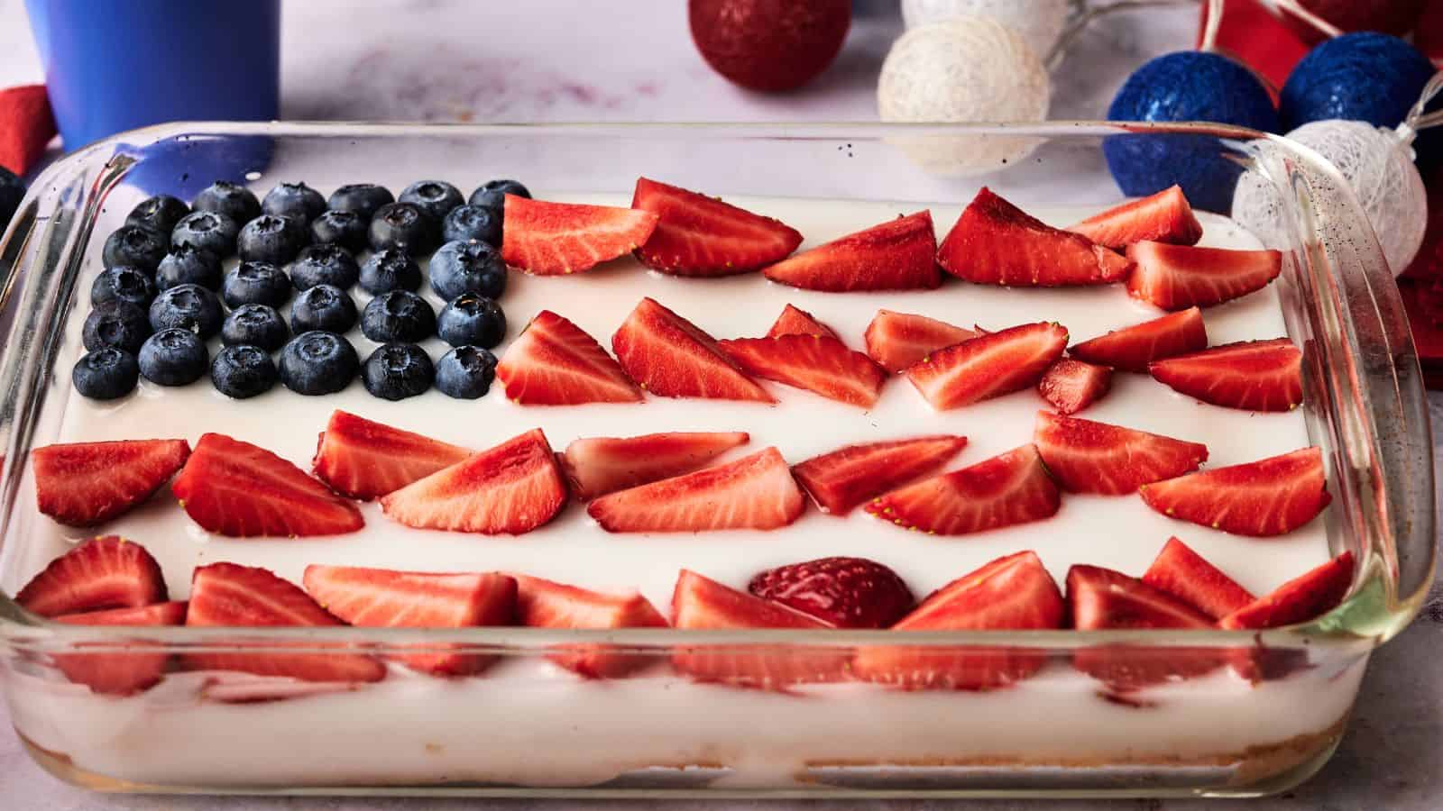 A rectangular dessert in a glass dish is topped with blueberries and sliced strawberries arranged to resemble the American flag.