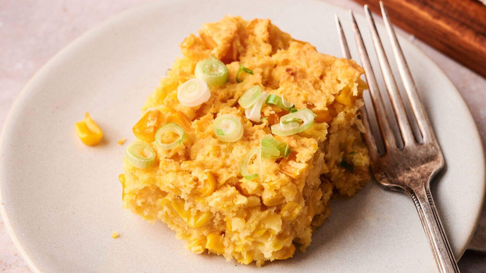 A square piece of cornbread with corn kernels and sliced green onions on top sits on a white plate next to a fork.