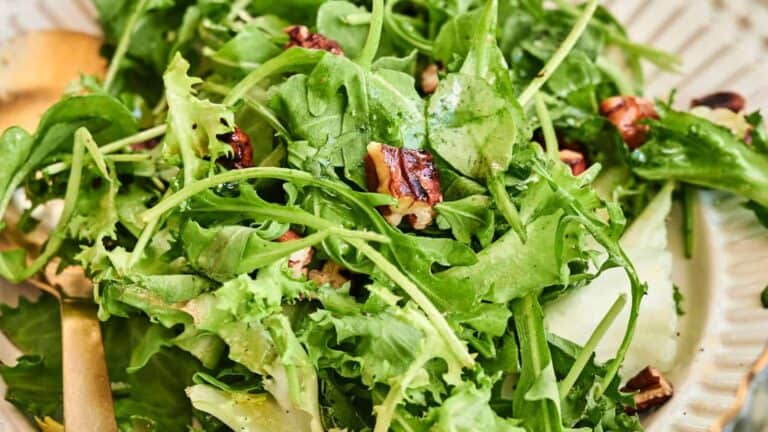 A close-up of a green salad with arugula, mixed leafy greens, pecan pieces, and a light dressing on a white plate.