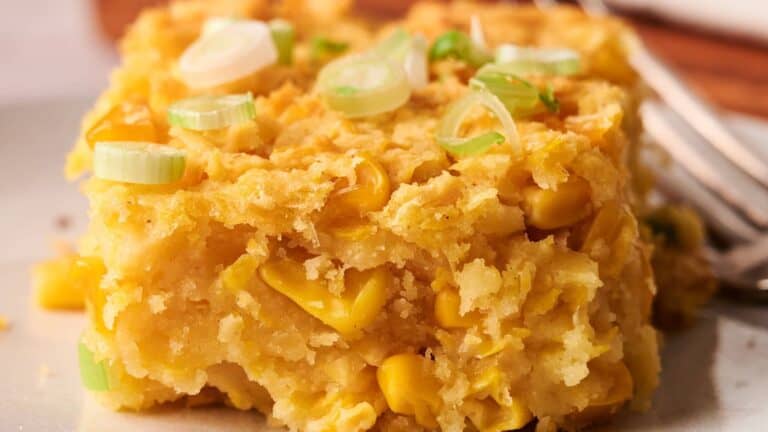 A square slice of corn casserole topped with chopped green onions, showing visible corn kernels and a moist, crumbly texture.