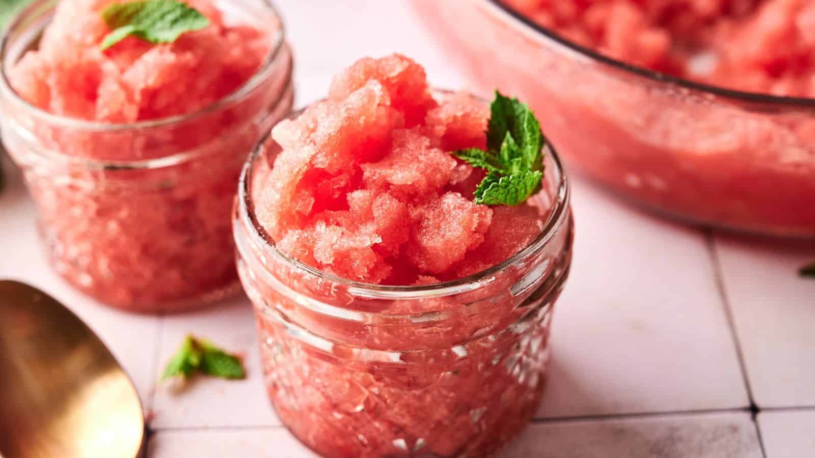 Two small glass jars filled with pink watermelon granita, garnished with mint leaves, are placed on a white tiled surface.