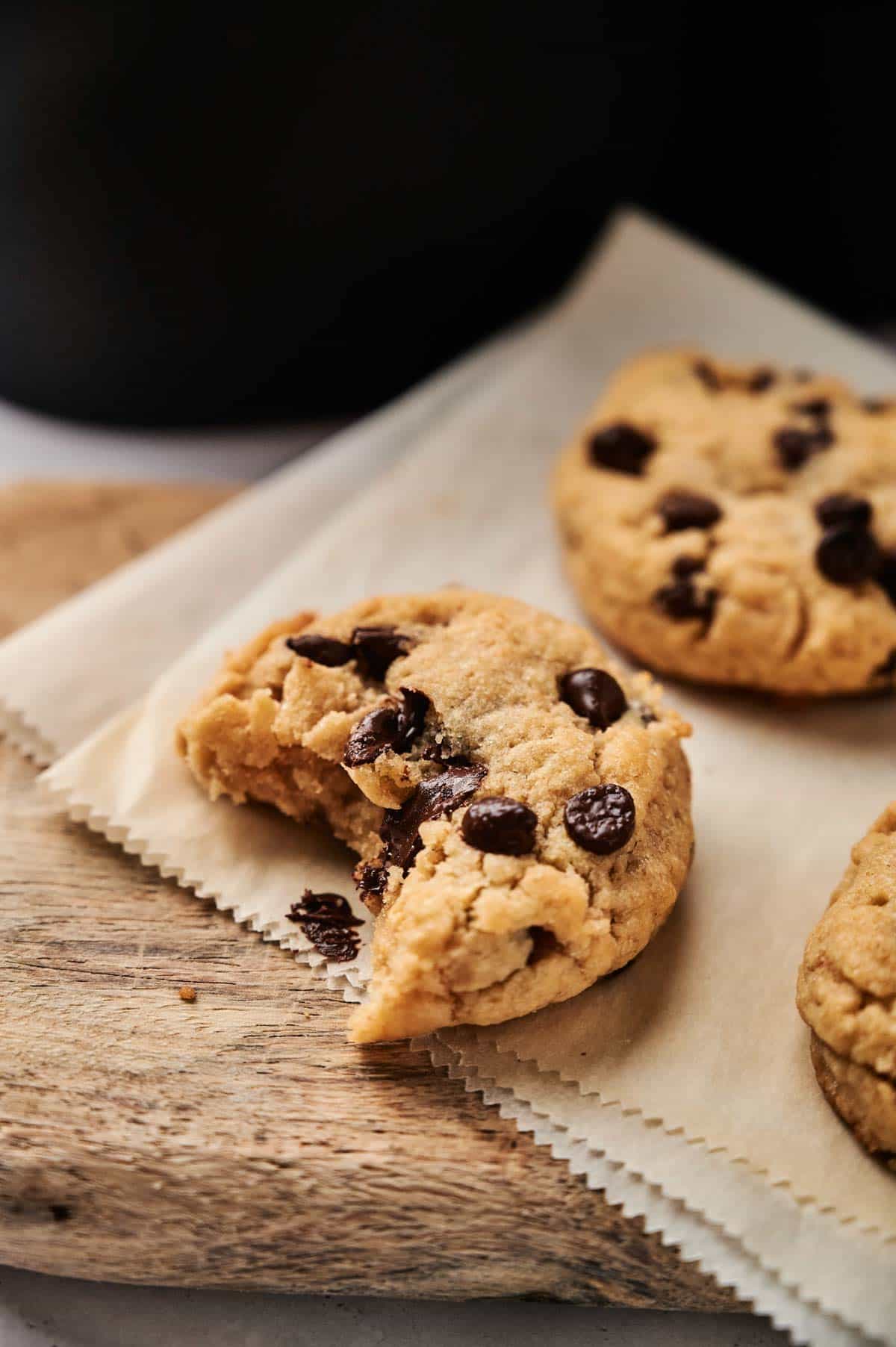 A close-up of an Air Fryer Vegan Chocolate Chip Cookie with a bite taken out of it, resting on parchment paper with more cookies in the background.