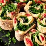 Vegan Pinwheels made with whole wheat tortillas, filled with hummus, spinach, black olives, and red peppers, then garnished with fresh parsley on a black plate.
