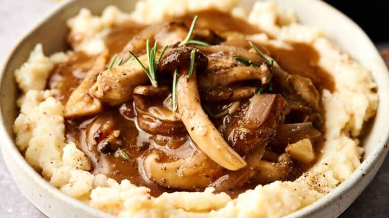 A bowl of mashed potatoes topped with mushroom and onion gravy, garnished with fresh rosemary.