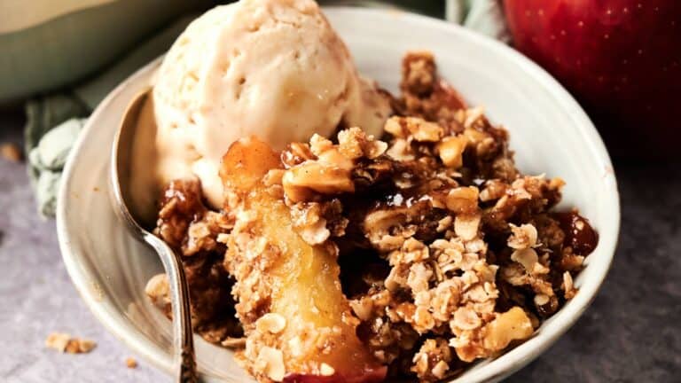A bowl of apple crisp topped with a scoop of vanilla ice cream, with a spoon placed in the bowl.