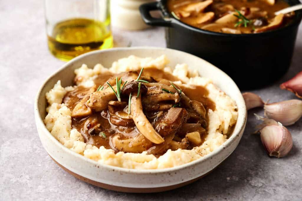 A bowl of mashed potatoes topped with vegan mushroom gravy and garnished with fresh rosemary; a pot of more gravy, garlic cloves, and a bottle of oil are in the background.