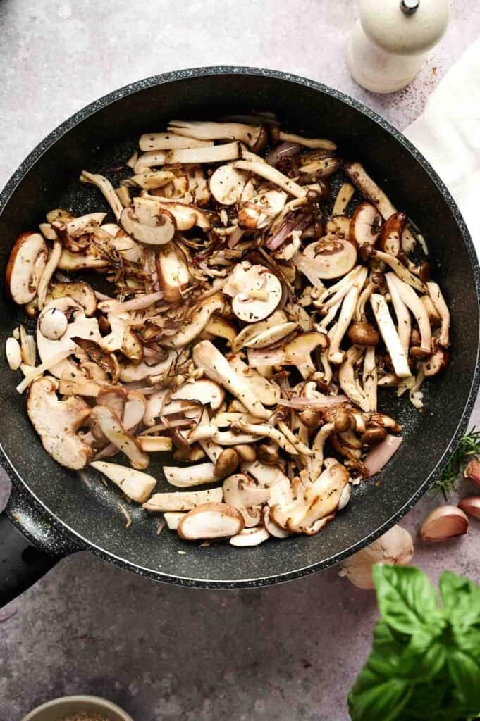 A frying pan filled with sliced mixed mushrooms and shallots being sautéed for a flavorful Vegan Mushroom Gravy, with garlic, herbs, and a basil leaf visible nearby.