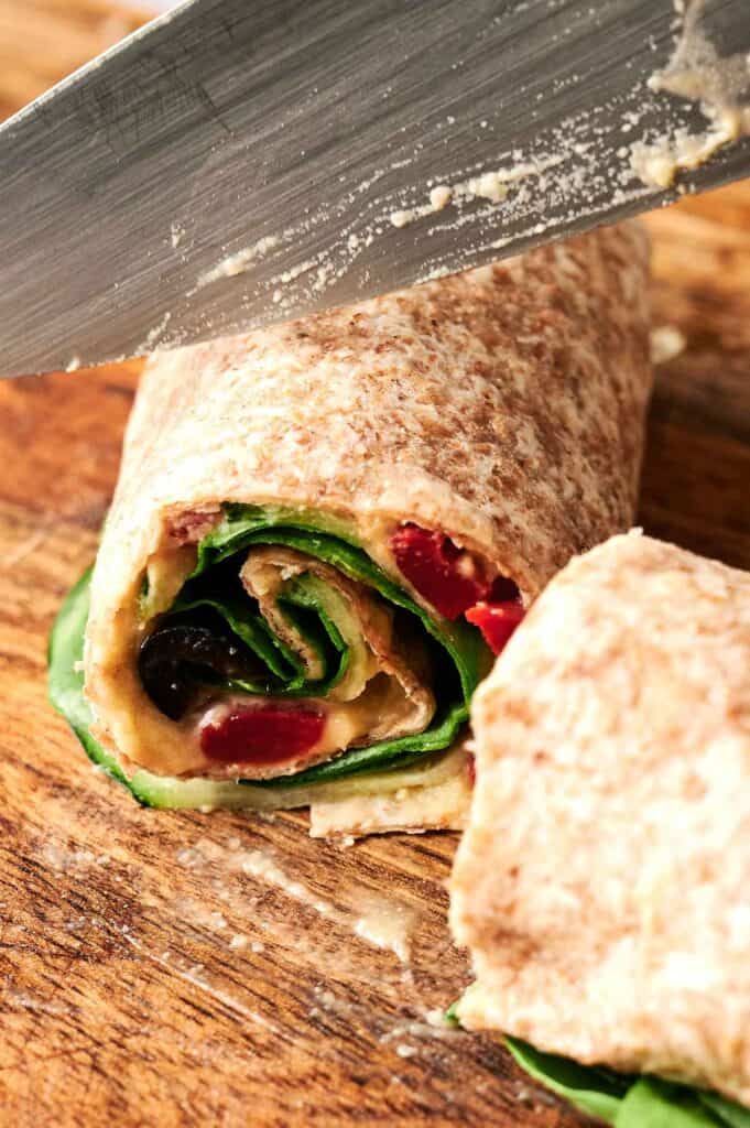 A close-up of Vegan Pinwheels: a whole grain wrap filled with spinach, hummus, and red peppers being sliced with a knife on a wooden surface.