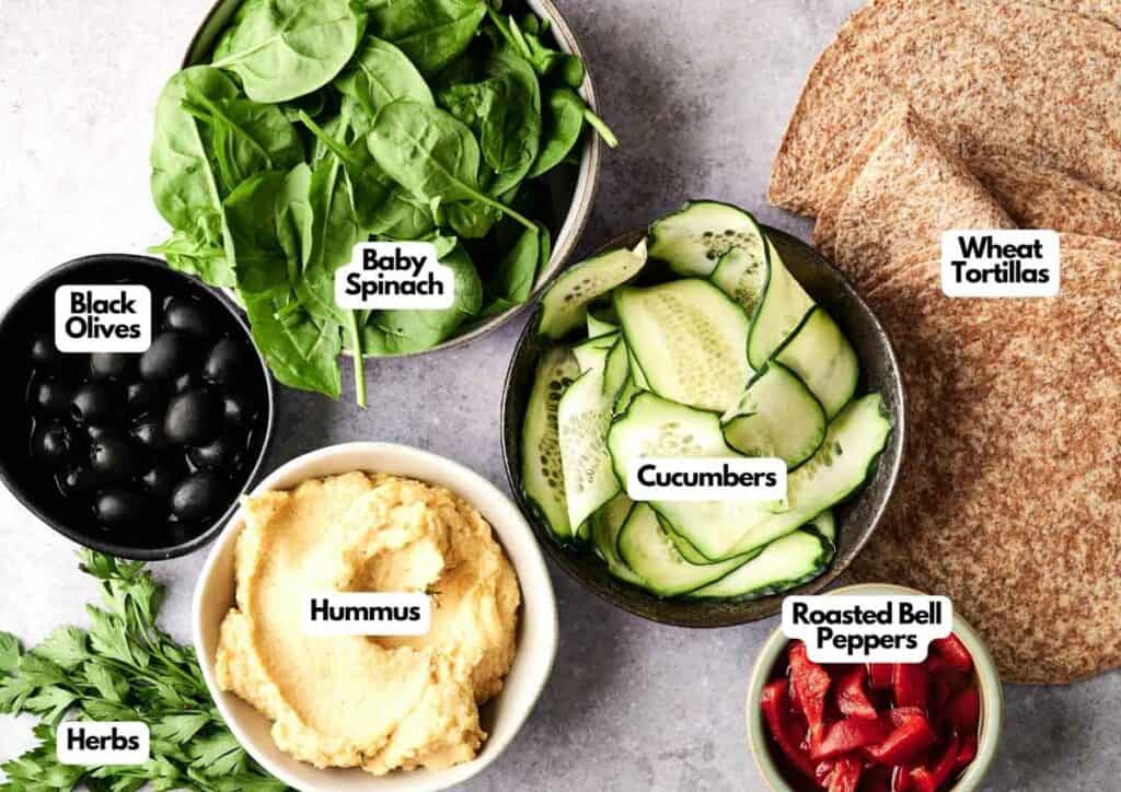 Bowls of black olives, hummus, cucumber slices, baby spinach, roasted bell peppers, fresh herbs, and wheat tortillas arranged on a countertop—perfect for assembling delicious Vegan Pinwheels.