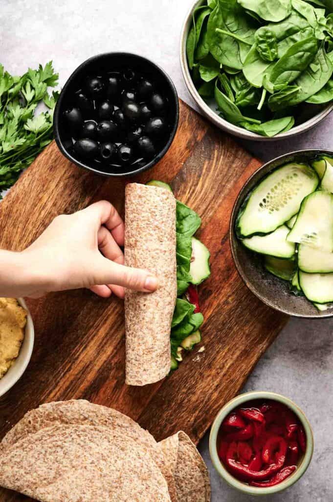 A hand rolls a whole wheat wrap filled with fresh spinach, cucumber, and other vegetables on a wooden board, creating delicious vegan pinwheels, surrounded by bowls of spinach, olives, cucumber, peppers, and herbs.