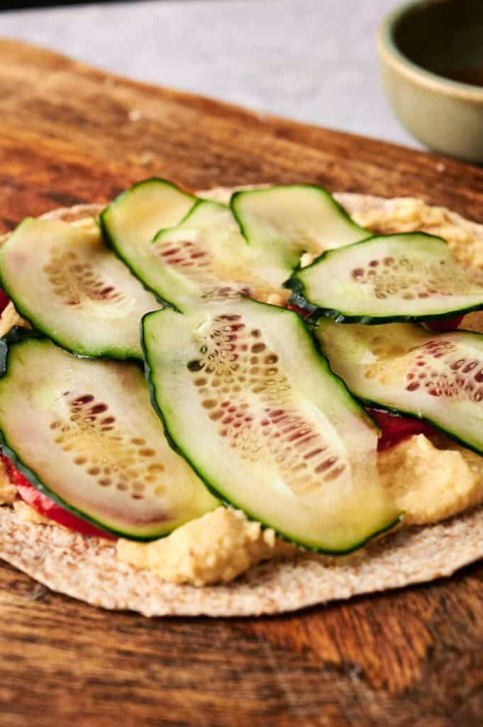 A whole wheat flatbread topped with hummus, sliced red peppers, and layers of cucumber slices on a wooden surface—perfect for rolling into delicious Vegan Pinwheels.