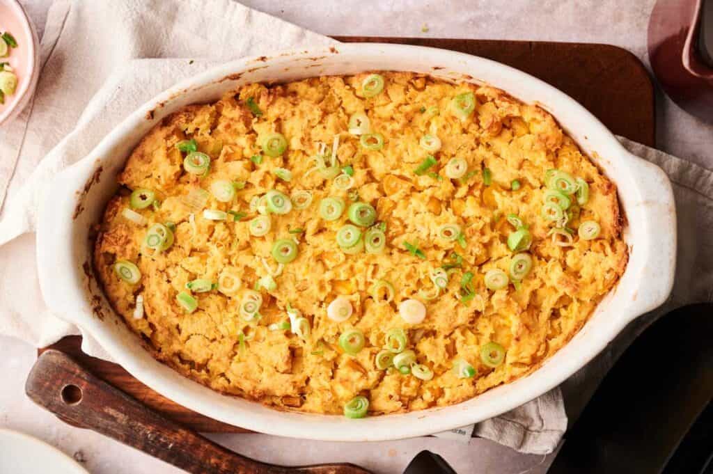 An Air Fryer Vegan Corn Casserole, baked to perfection and topped with sliced green onions in an oval white dish, sits on a wooden board and a beige cloth.