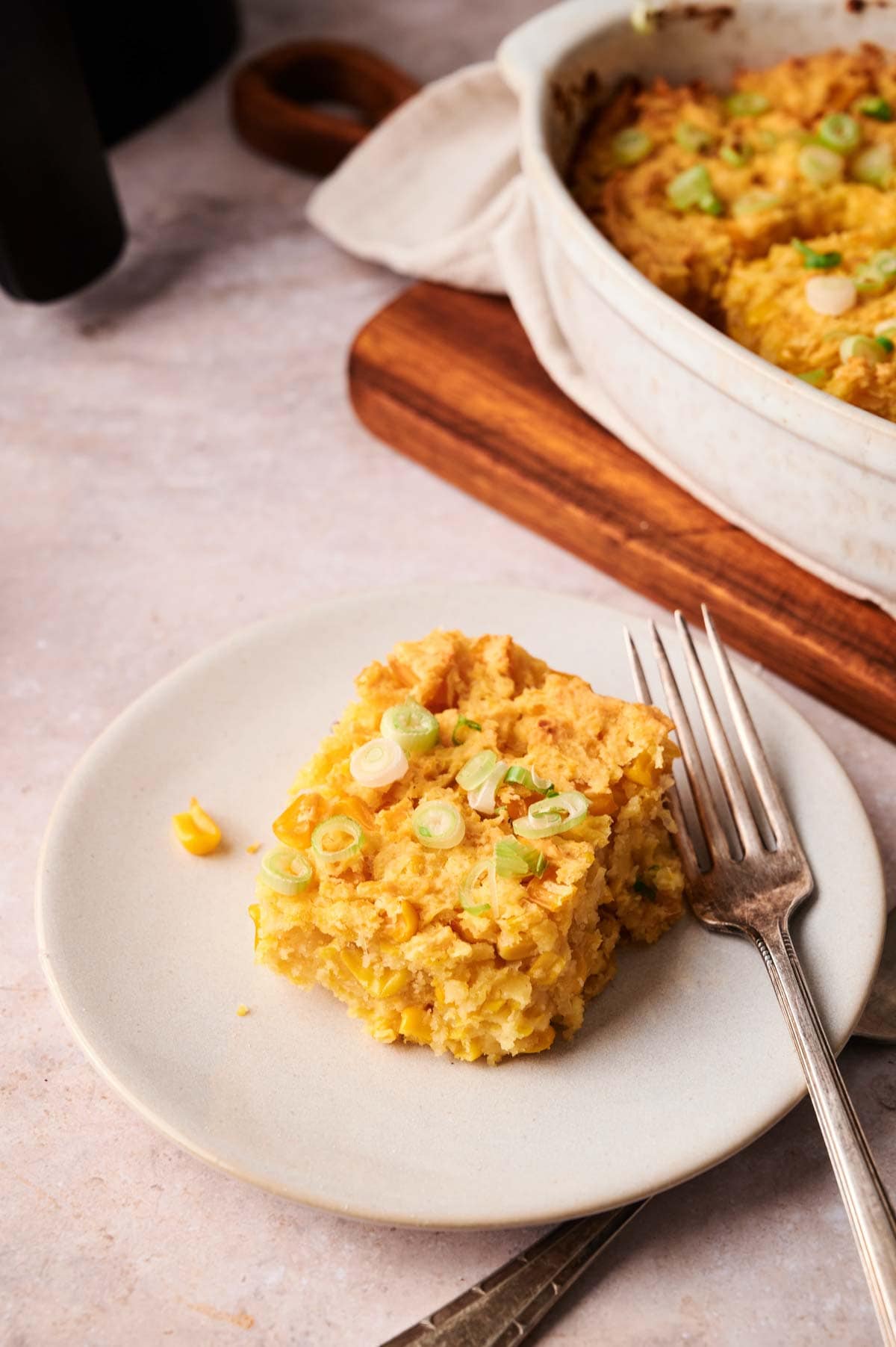 A square serving of Air Fryer Vegan Corn Casserole topped with chopped green onions on a white plate, with a fork beside it and a baking dish in the background.