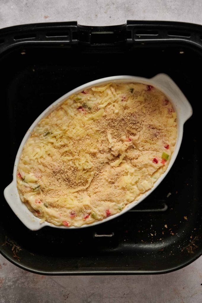 An oval baking dish filled with Air Fryer Vegan Hashbrown Casserole, topped with grated cheese and breadcrumbs, sits inside an air fryer basket.