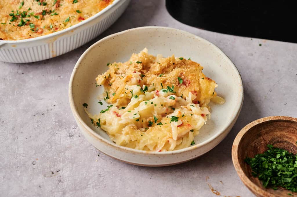 A serving of baked cheesy casserole, reminiscent of Air Fryer Vegan Hashbrown Casserole, topped with breadcrumbs and chopped parsley on a beige plate, with a casserole dish and a bowl of parsley nearby.