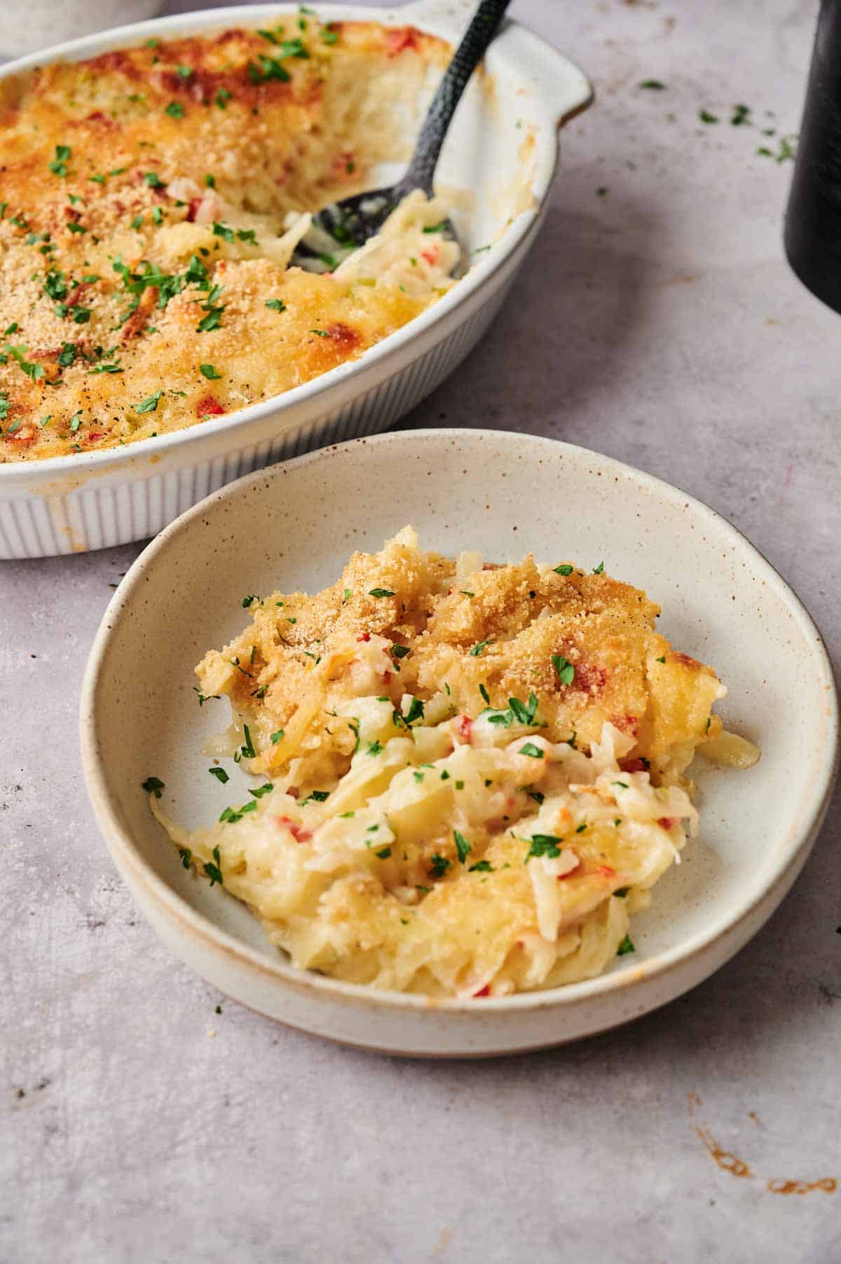 A serving of cheesy Air Fryer Vegan Hashbrown Casserole topped with breadcrumbs and chopped herbs is plated in a bowl, with the rest of the casserole in a baking dish in the background.