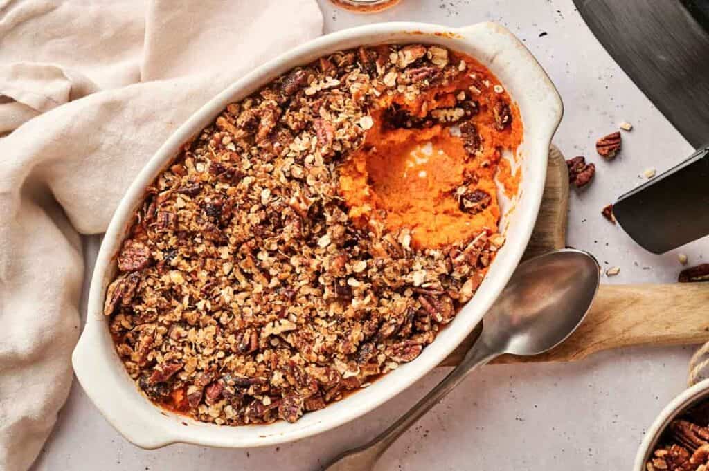 An oval baking dish filled with Air Fryer Sweet Potato Casserole, topped with pecan and oat crumble, sits with a serving missing. A spoon and napkin are nearby.