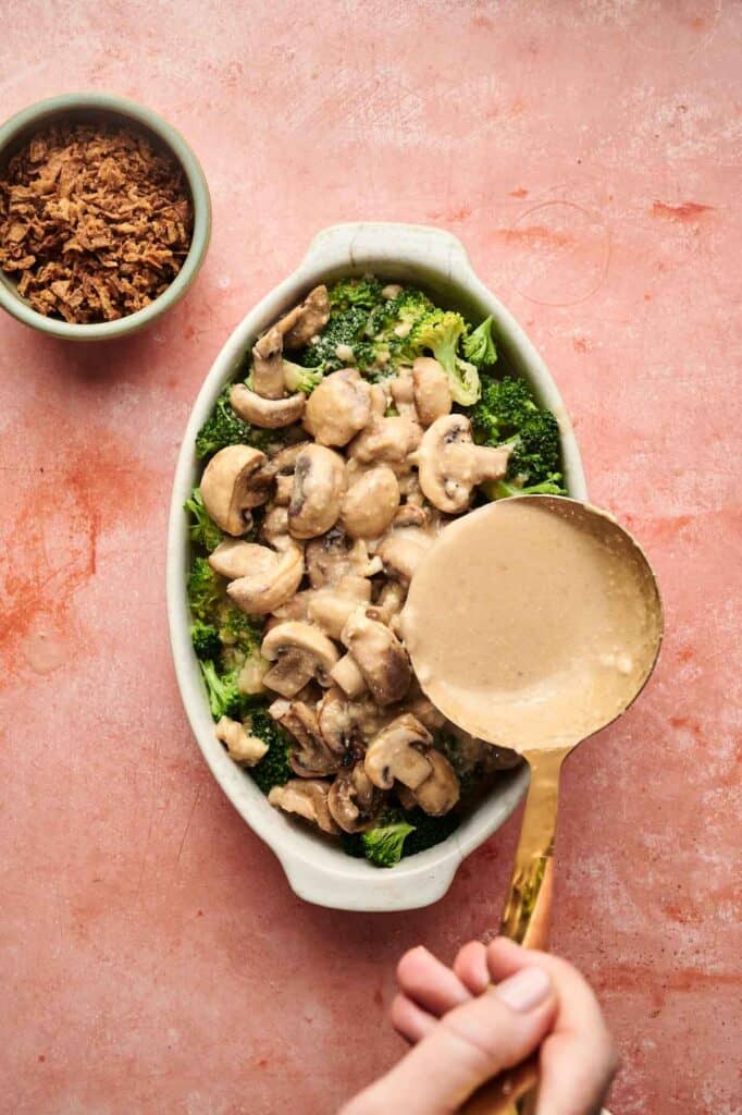 A hand pours creamy mushroom sauce over broccoli and mushrooms in a white baking dish, creating a delicious Vegan Broccoli Casserole, with a small bowl of crispy onions beside it on a pink surface.
