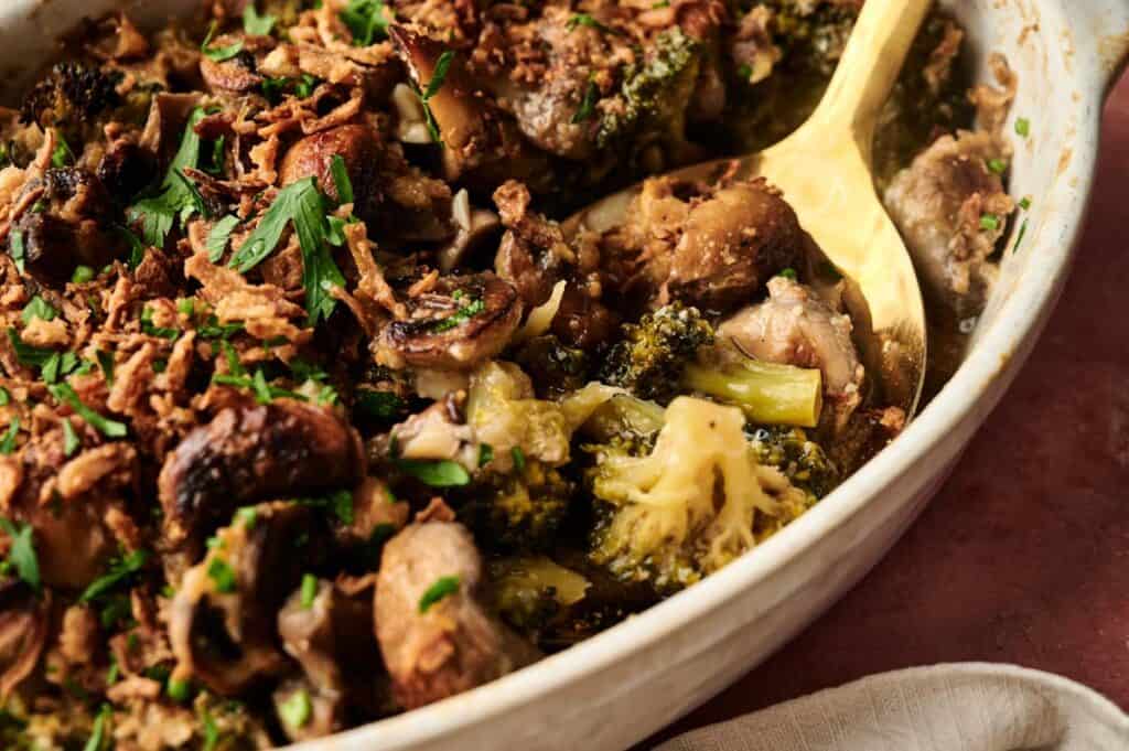 A close-up of a Vegan Broccoli Casserole with mushrooms, crispy onions, and herbs, served in a ceramic dish with a wooden spoon.