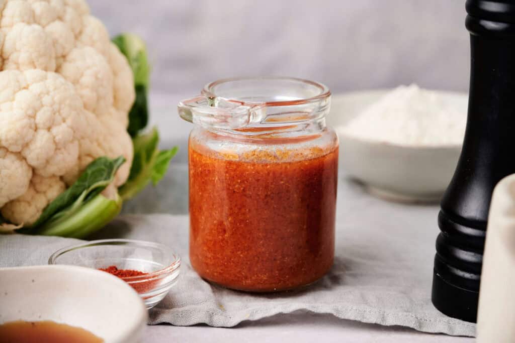 A glass jar of reddish-brown Buffalo Sauce sits on a cloth, surrounded by a cauliflower, a bowl of flour, a pepper grinder, and a small bowl of paprika.