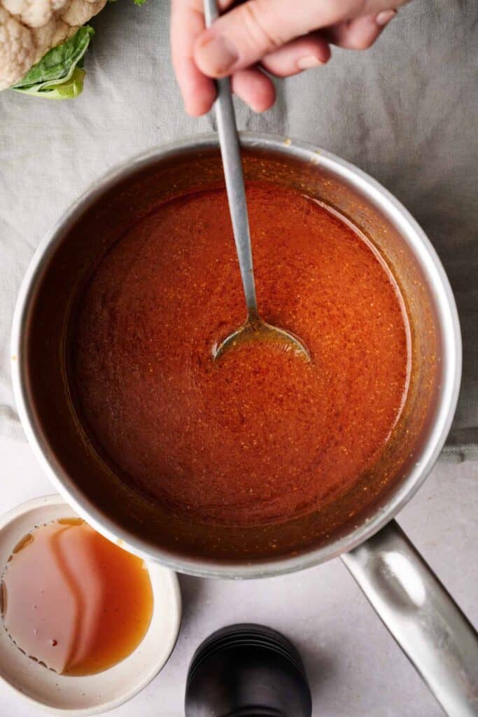 A hand stirs a pot of reddish-brown Buffalo Sauce with a spoon; a small dish of liquid and part of a cauliflower are nearby on a gray cloth.