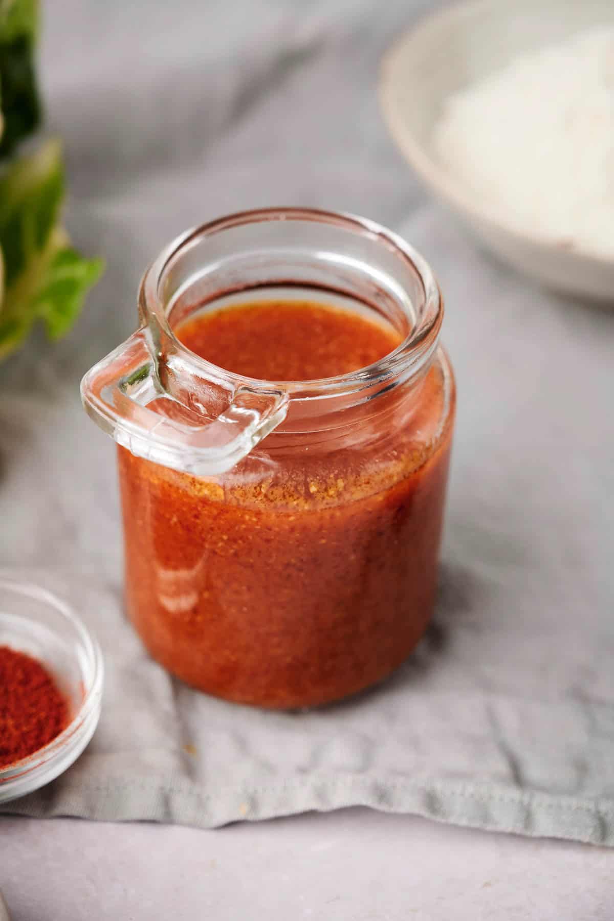 A small glass jar filled with spicy red chili paste, reminiscent of Buffalo Sauce, sits on a gray cloth, with a small bowl of red powder and a white bowl in the background.