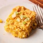A square piece of Air Fryer Vegan Corn Casserole with corn kernels and chopped green onions on top, served on a white plate with a fork.