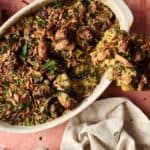 A Vegan Broccoli Casserole in a white dish is topped with crispy onions and fresh herbs, with a serving spoon lifting out a portion. A beige napkin is placed beside the casserole, ready to serve.