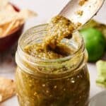 A spoon scoops Salsa Verde from a glass jar, with tortilla chips and a tomatillo in the background.
