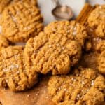 A batch of Vegan Peanut Butter Cookies sprinkled with coarse salt is arranged on a wooden board, with a plate and spoon in the background.