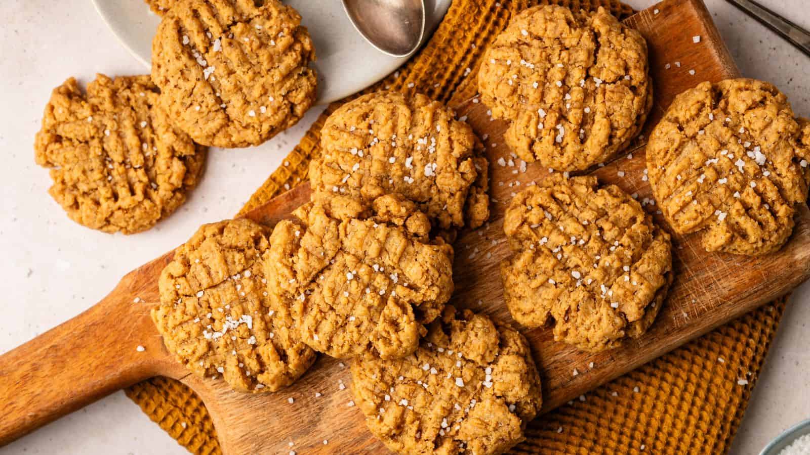 A wooden board with several peanut butter cookies topped with coarse salt, next to a plate with more cookies and a metal spoon.