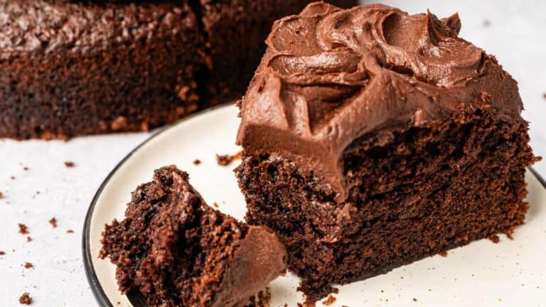 A slice of chocolate cake with thick chocolate frosting on a white plate, with a bite-sized piece and crumbs beside it.