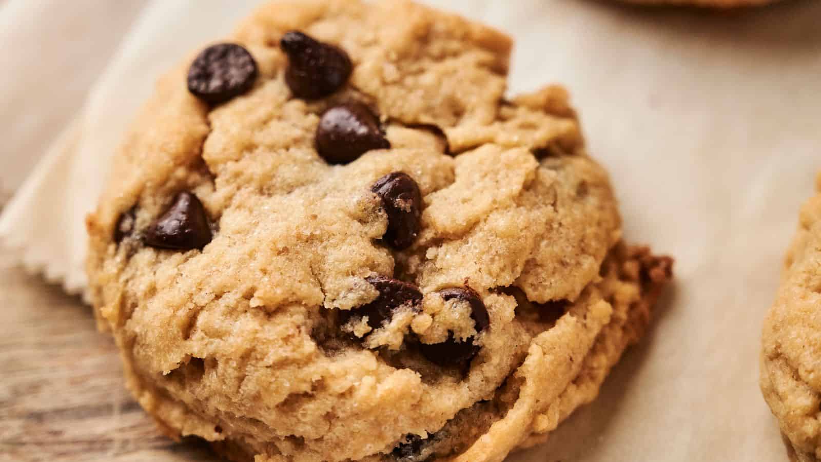A close-up of a chocolate chip cookie resting on parchment paper, showing visible chocolate chips and a soft, crumbly texture.