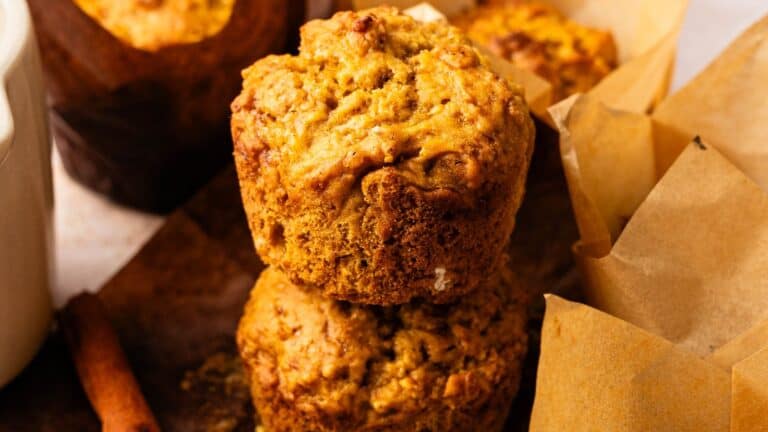 Two pumpkin muffins stacked on a wooden surface, with more muffins wrapped in parchment paper in the background.