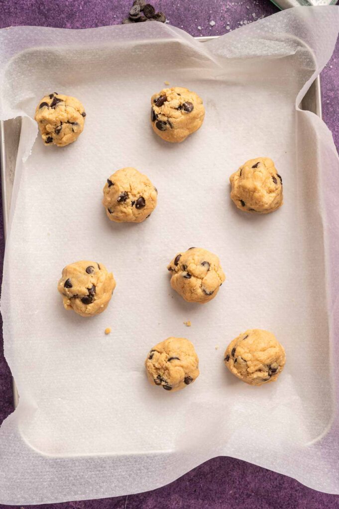 Seven chocolate chip cookie dough balls arranged on a parchment-lined baking sheet, ready to be baked.