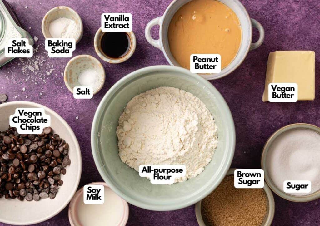 Various baking ingredients in bowls on a purple surface, including labeled peanut butter, vegan butter, all-purpose flour, brown sugar, sugar, soy milk, vegan chocolate chips, salt, and vanilla extract.