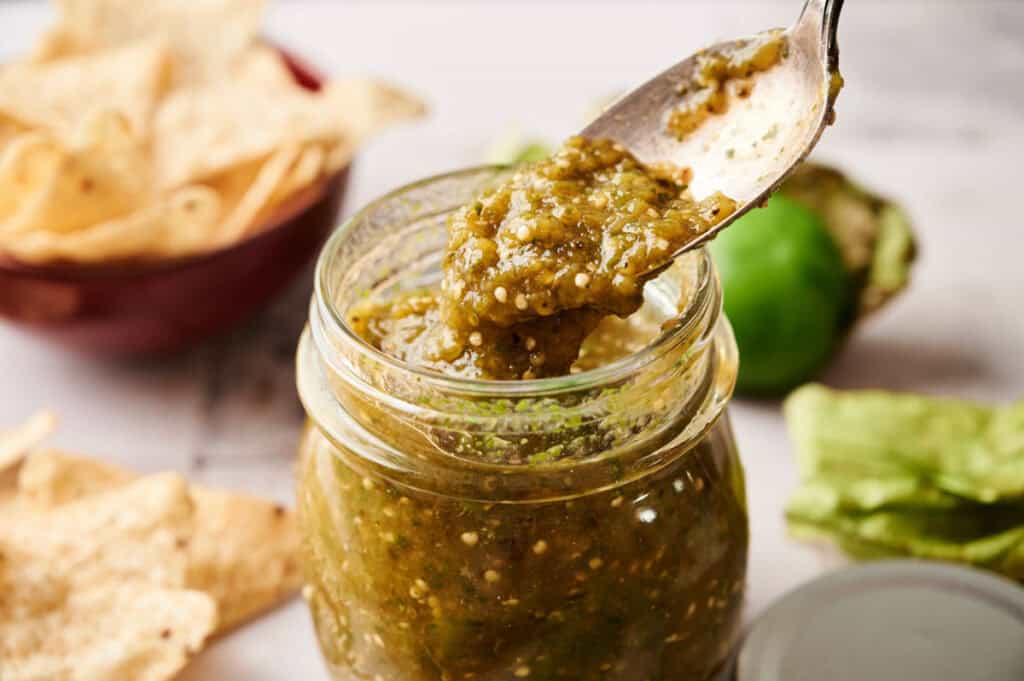 A spoon scooping vibrant Salsa Verde from a glass jar, with tortilla chips and tomatillo ingredients in the background.
