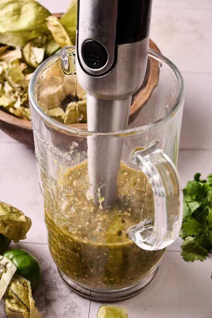 An immersion blender blends Salsa Verde ingredients in a glass measuring cup, with tomatillos, cilantro, and lime nearby on a light countertop.