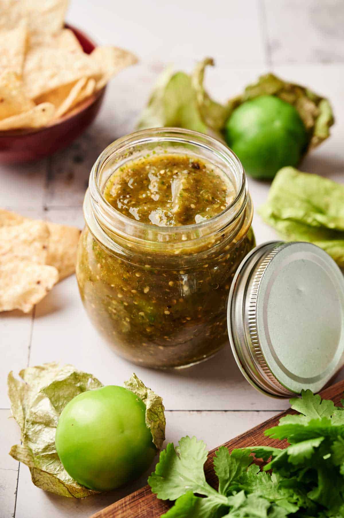A jar of Salsa Verde sits open on a table with tomatillos, cilantro, and tortilla chips nearby.