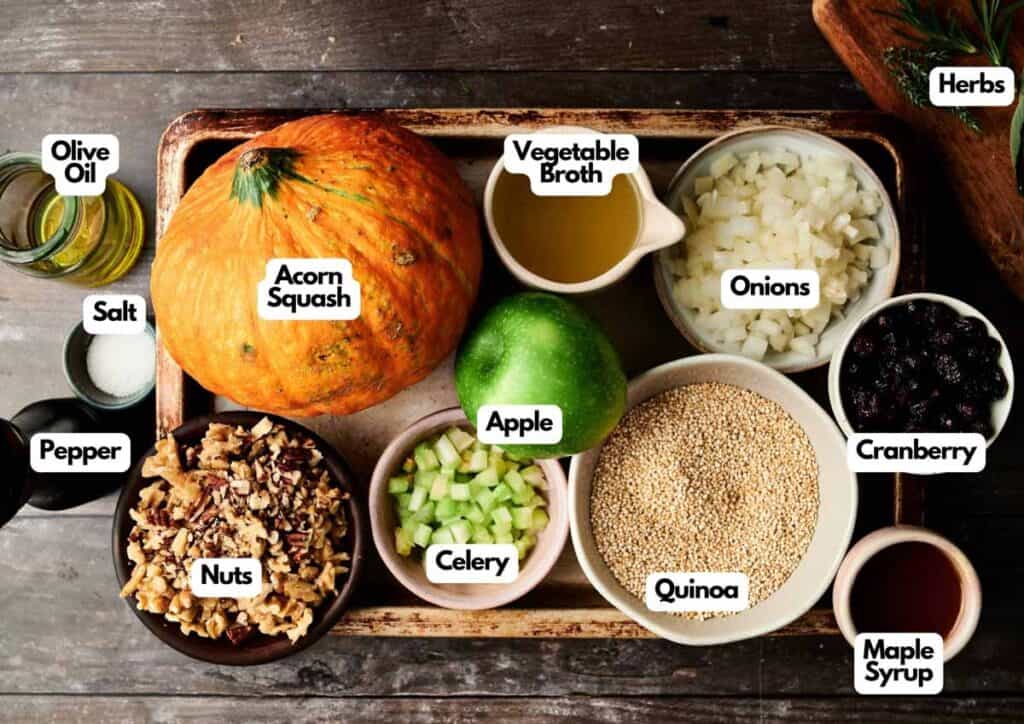A wooden tray holds bowls and jars of ingredients for Stuffed Acorn Squash, including apple, celery, onion, quinoa, nuts, dried cranberries, herbs, olive oil, vegetable broth, salt, and pepper.