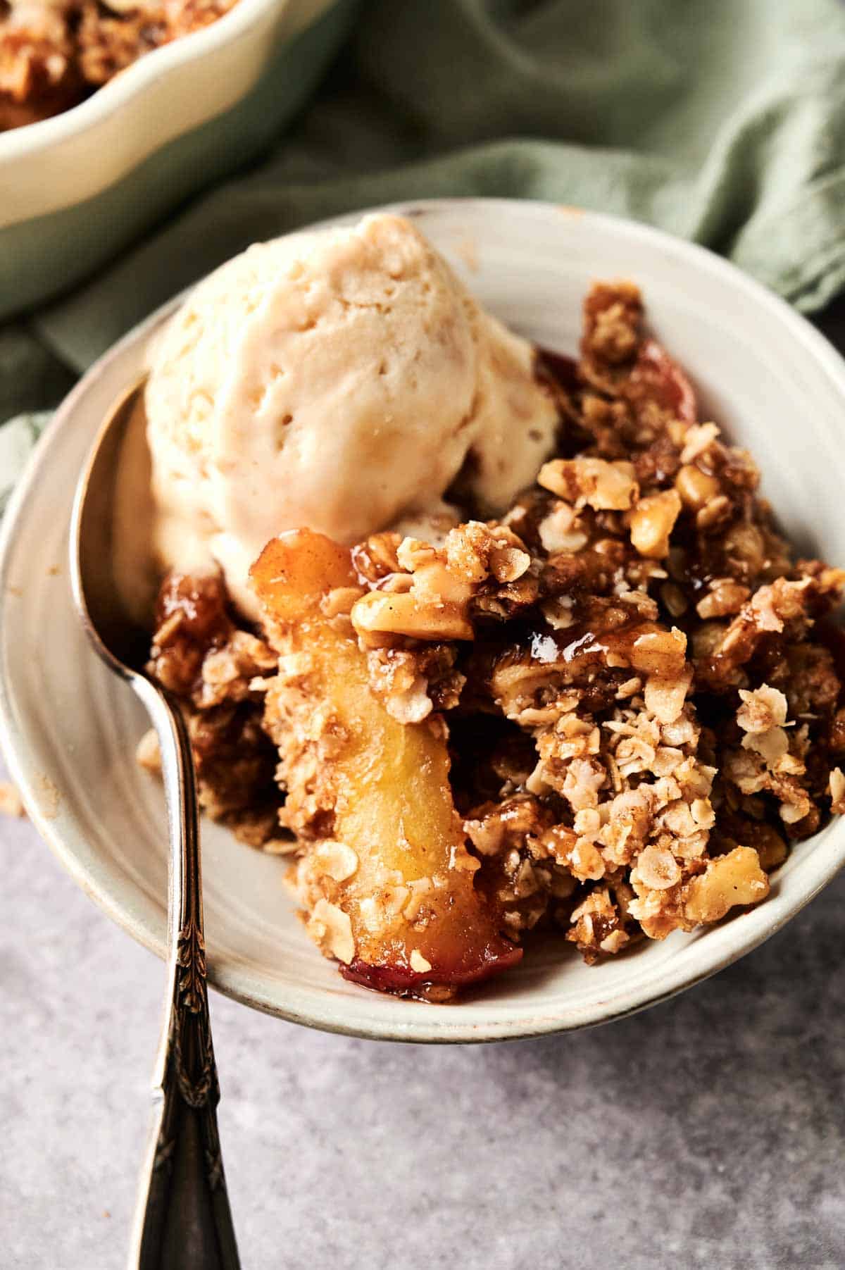 A bowl of peach crisp topped with a scoop of vanilla ice cream, served with a spoon&mdash;perfect for anyone who loves Vegan Apple Crisp and sweet, comforting desserts.