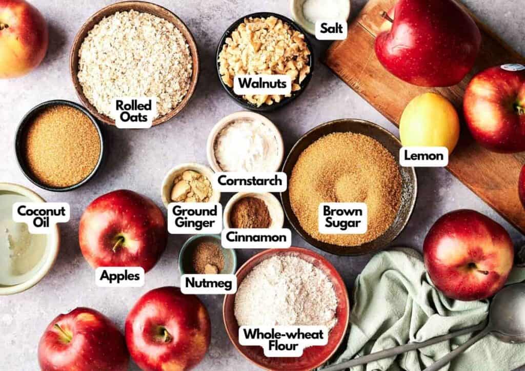 Top-down view of ingredients for Vegan Apple Crisp: apples, brown sugar, whole-wheat flour, rolled oats, walnuts, coconut oil, ground ginger, nutmeg, cinnamon, lemon, cornstarch, and salt arranged on a counter.
