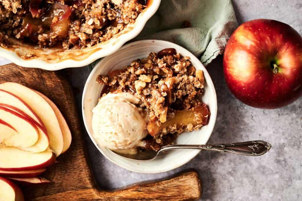A bowl of Vegan Apple Crisp with a scoop of vanilla ice cream sits next to a whole apple, sliced apples, and a baking dish on a gray countertop.