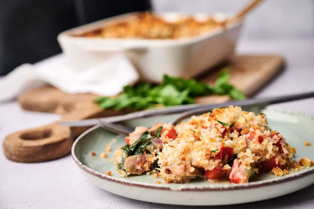 A serving of Air Fryer Vegan Breakfast Casserole topped with breadcrumbs is on a plate with a fork, while the rest of the casserole sits in a baking dish in the background.