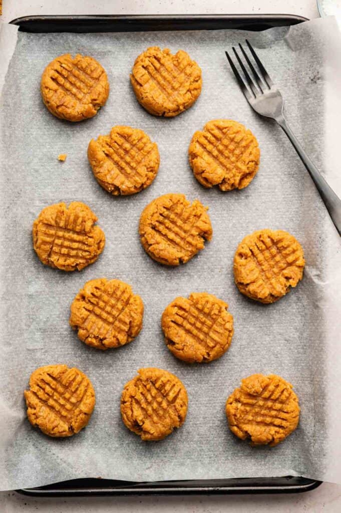 A baking tray lined with parchment paper holds twelve unbaked Vegan Peanut Butter Cookies with a crisscross fork pattern on top; a fork rests beside them.