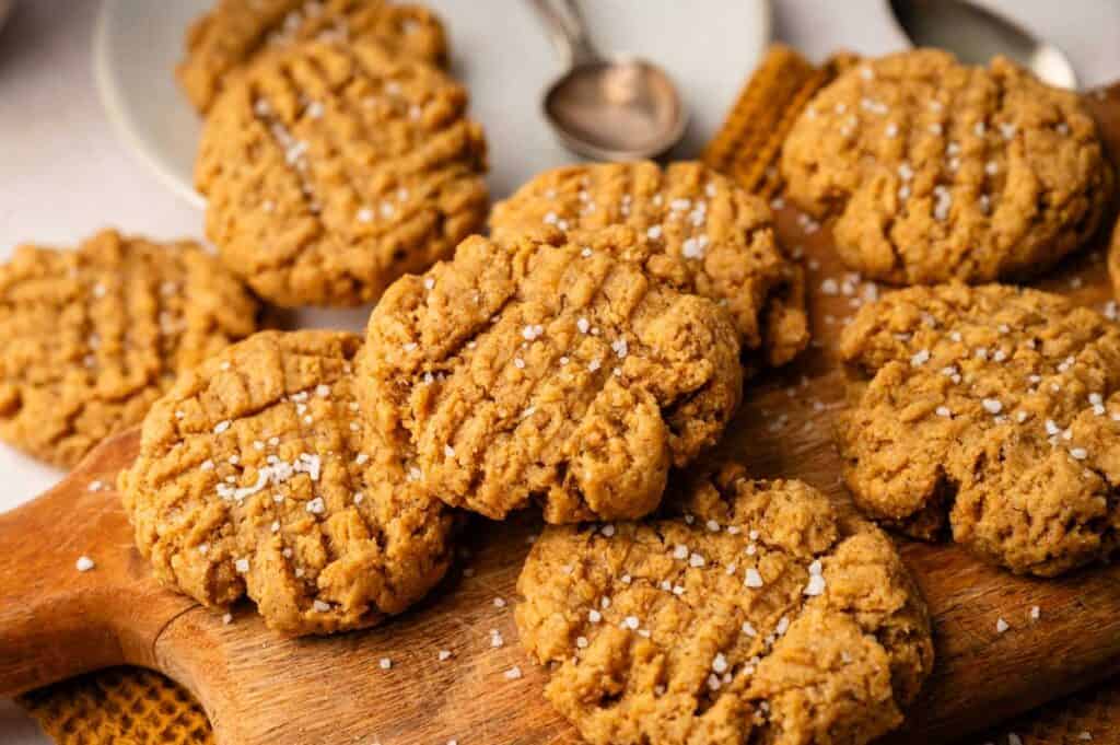 A wooden board with several Vegan Peanut Butter Cookies topped with coarse salt, with plates and a spoon in the background.