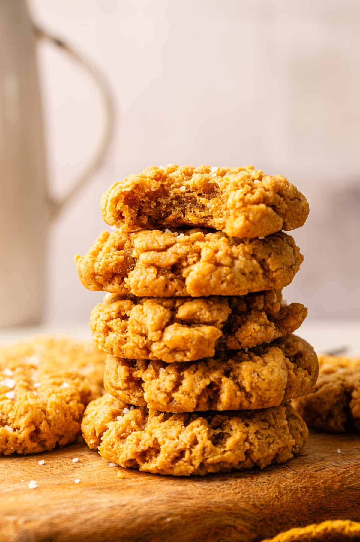 A stack of five Vegan Peanut Butter Cookies, with a bite taken out of the top cookie, rests on a wooden surface.