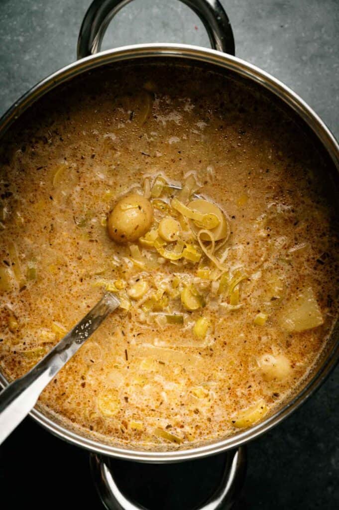 A pot of creamy Vegan Potato Soup with leeks and spices sits on a dark surface, a metal ladle partially submerged in the hearty soup.